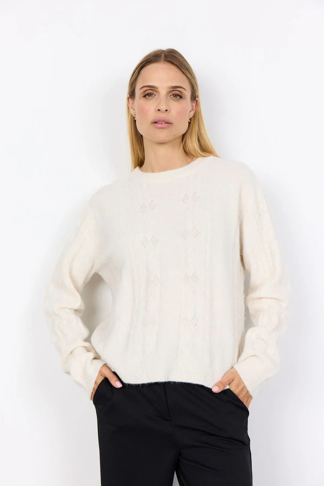 Cremeweißer Strickpullover mit feinem Ajourmuster, kombiniert mit einer schwarzen Hose, vor hellem Studiohintergrund.