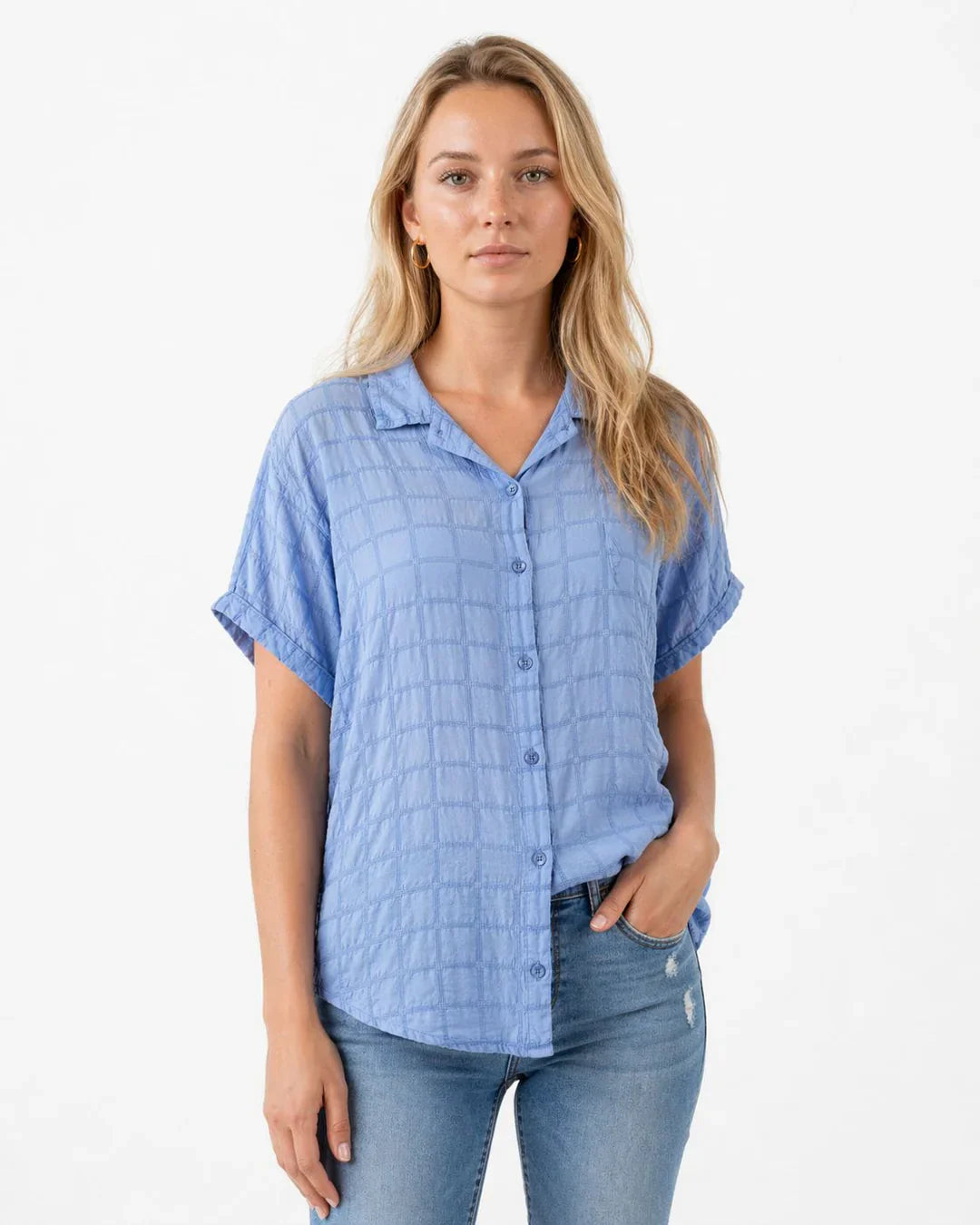 Frau in hellblauer Kurzarmbluse mit feiner Struktur und lässiger Jeans vor schlichtem Studiohintergrund. Der leichte Look kombiniert sommerliche Eleganz mit modernem Casual-Style für Alltag und Freizeit.