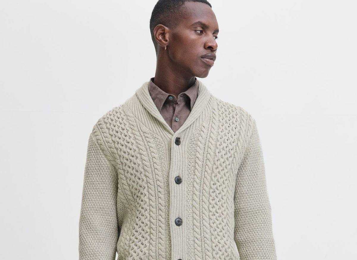 Herren-Strickjacke in Creme mit markantem Strickmuster und dunklen Knöpfen, kombiniert mit Hemd und heller Jeans im modernen Studio-Look.