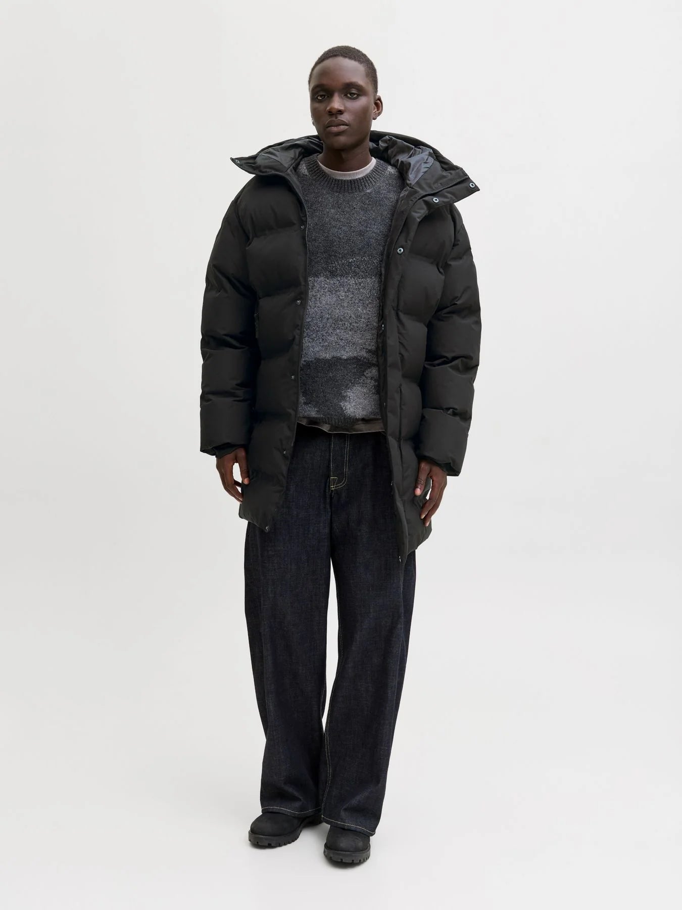 Schwarzer Herren-Steppmantel über grauem Strickpullover und dunkler Jeans, kombiniert mit schwarzen Boots vor hellem Studiohintergrund.
