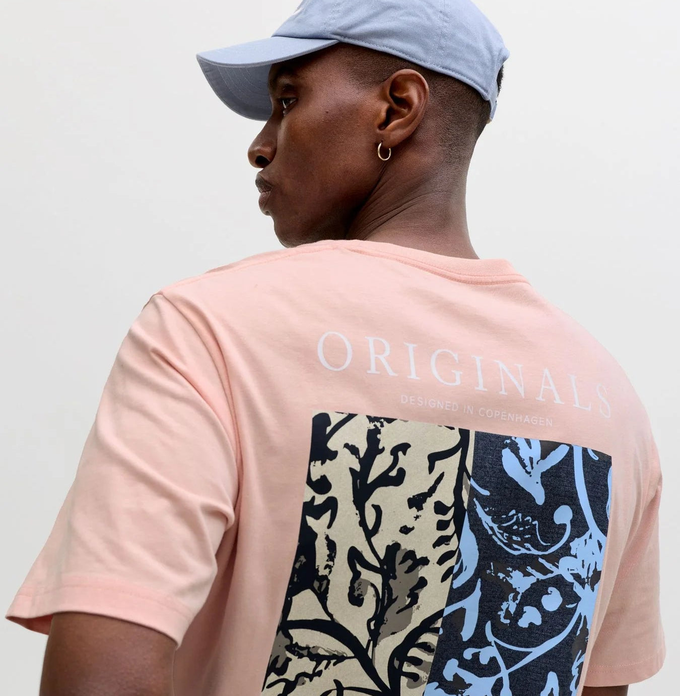 Rosa Herren-T-Shirt mit großem Print auf der Rückseite, getragen mit hellblauer Cap vor hellem Studiohintergrund.