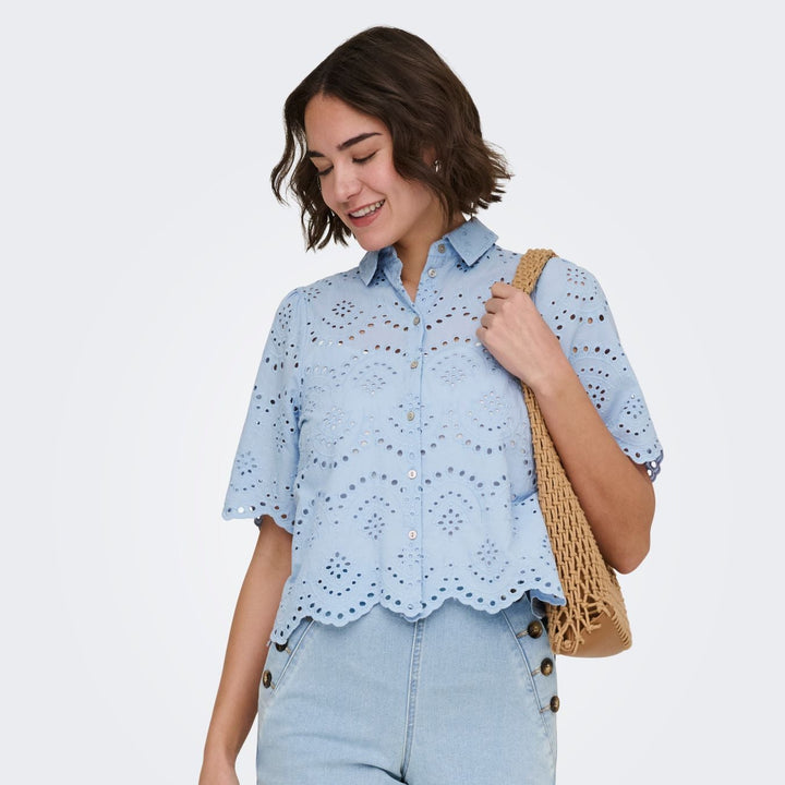Junge Dame mit kurzen braunen harren trägt Bluse mit Lochmuster in Pastellblau und Makramee Tasche 