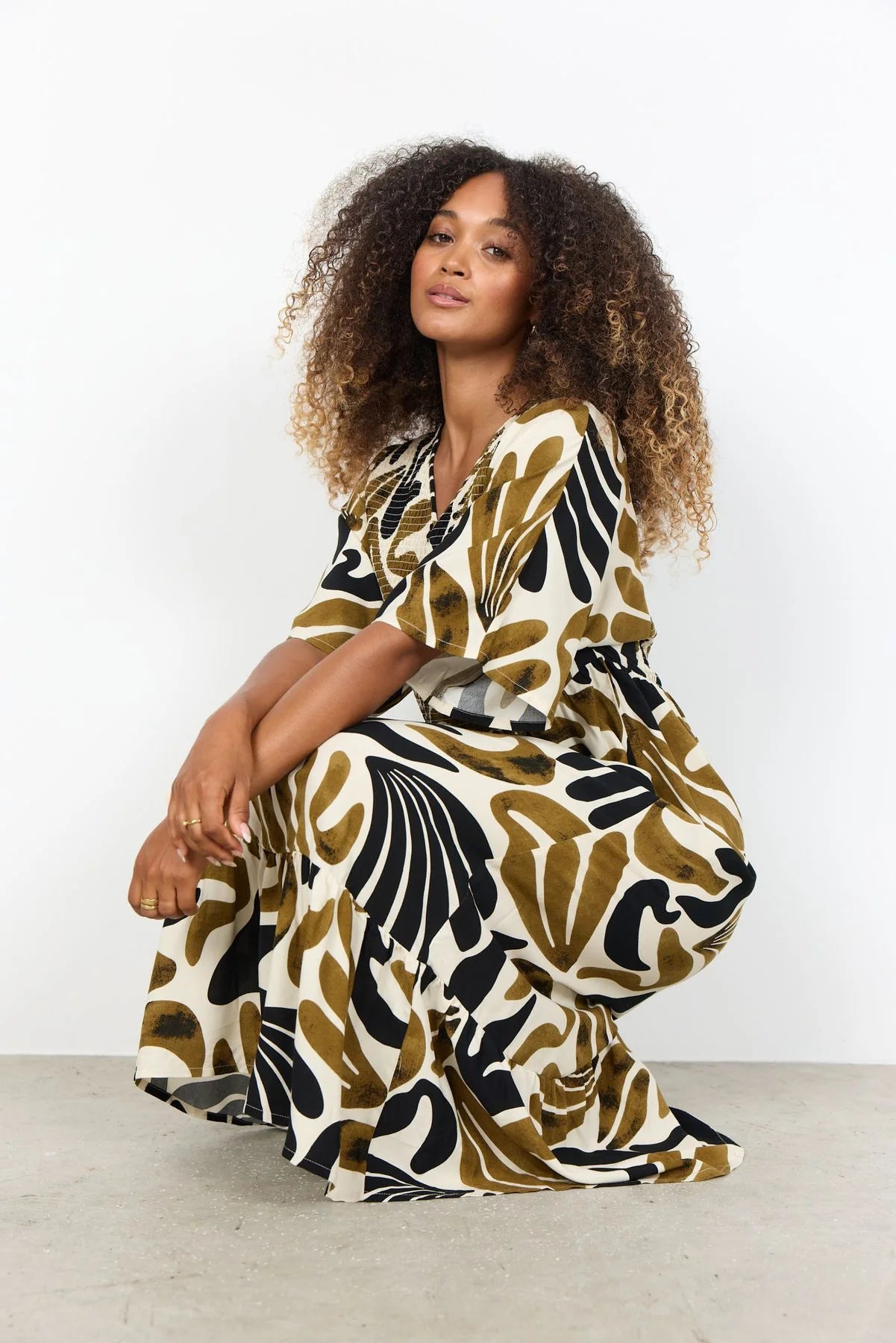 Gemustertes Maxikleid in Creme, Schwarz und Oliv, getragen von einem sitzenden Model vor hellem Studiohintergrund.
